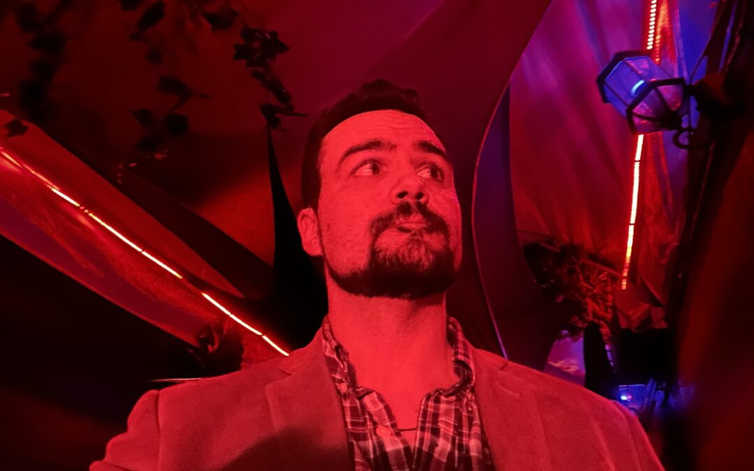 40-year old white man with dark hair, dark eyes, and a dark chinstrap beard, wearing a plaid shirt and corduroy jacket standing in a dark hallway, bathed in neon red light