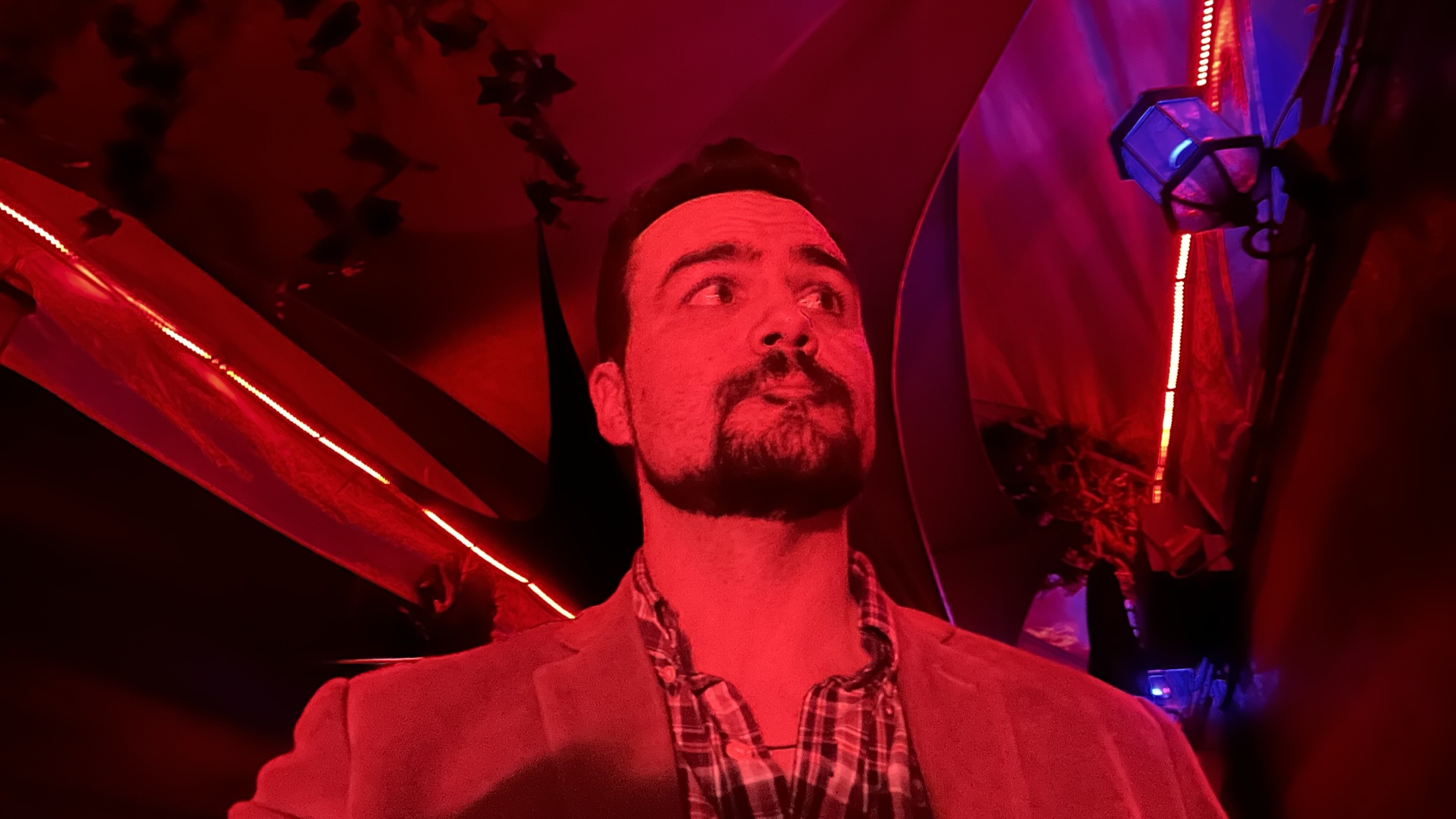 40-year old white man with dark hair, dark eyes, and a dark chinstrap beard, wearing a plaid shirt and corduroy jacket standing in a dark hallway, bathed in neon red light