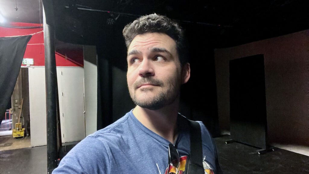 A selfie of Gregory Crafts, standing on stage, half-cloaked in shadow, and looking wistfully around his venue, studio/stage. Enjoying the calm before the storm.