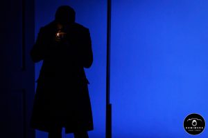 Silhouette of a man standing in a door frame, lighting a cigarette, standing against a blue background